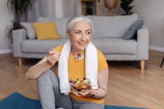 Image Happy senior woman eating fresh vegetable salad, sitting on yoga mat after home workout