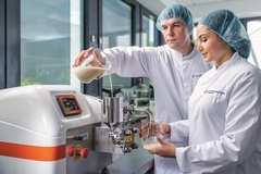 two technologists in lab coats pouring light brown thick liquid into large steel equipment