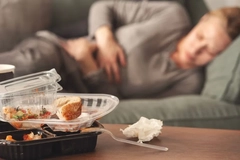 Image A woman clutches her stomach while lying on the couch with empty cartons of take out food on the table in front of her.