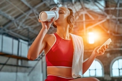 Image woman using smartphone and listening to music in headphones drinks protein shake while exercising in the fitness room.