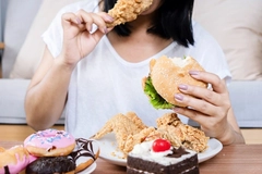 Image woman in white shirt biting into fried chicken with burger and donuts on table