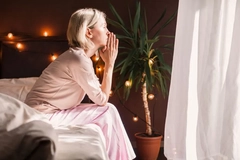 Image A woman sitting on the bed with her hands on her chin looking out a window.