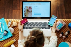 Image a person online shopping, holding phone and credit card in front of open laptop with cosmetic products scattered around.