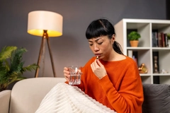 Image woman in orange sweater sitting on sofa holding a glass of water and a capsule