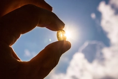 Image Hand holding soft gel fish oil or vitamin d capsule against blue sky and sun.