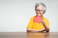 Image An elderly Asian woman is holding a brain symbol smile and looking at the camera while sitting at the table.