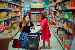 Image Mother Saying No to Child putting sweets in shopping basket. Mom gesturing no as her kid tries adding sugary snacks