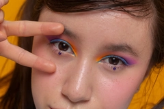 Image A girl wearing colorful eye makeup and doing a peace sign in front of her eye.