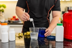 Image Sportsman pours scoop of supplement powder into glass of water in home kitchen.
