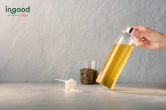 Image Hand holding a glass bottle of golden oil beside a scoop of white powder and a glass of seeds on a neutral surface.