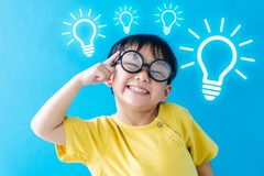 boy wearing glasses, smiling points his index finger to his head, with light bulbs above his head