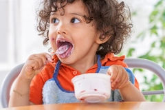 child licks spoon of yogurt, holding a bowl at dining table