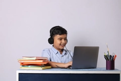 Image Indian boy sitting on desk with books, studying online using laptop.