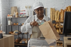 Image woman working in a bakery, packing a pastry into a brown paper bag in a shop with various bread and pastries behind her