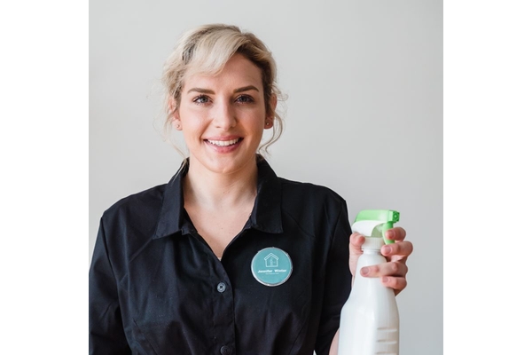 smiling woman in cleaners uniform with a badge on her breast holds a cleaning spray in her right hand.