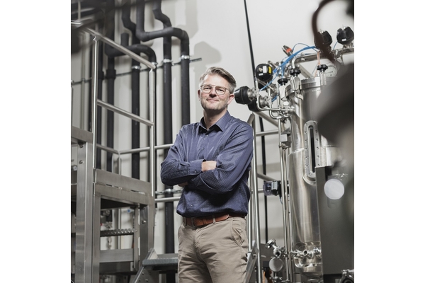 A man with glasses in a dark blue shirt and beige pants standing with crossed arms in a food factory with machinery behind him.