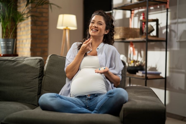 A pregnant women smiles while sitting on the couch and preparing to take a supplement.