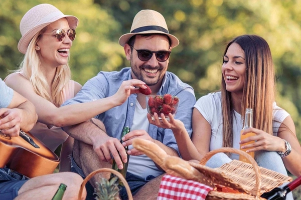 Image two young women and a man smiling and sharing strawberries in a park