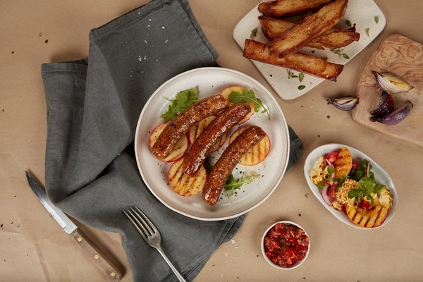 cultivated meat sausages on a white plate next to grey table cloth and dips
