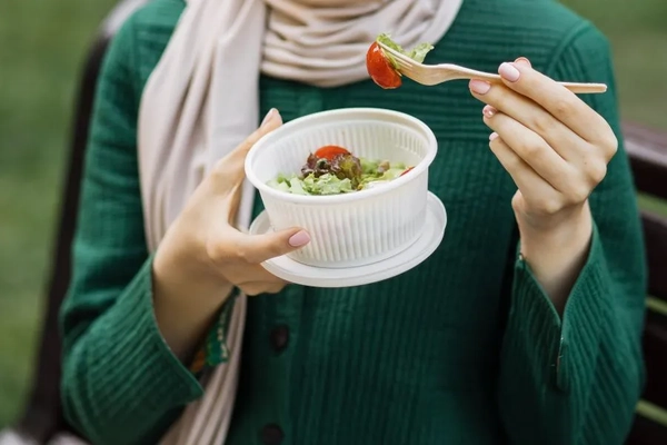 A person holding a plastic bowl of salad and eating it with a plastic fork