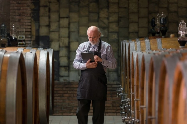 A winery expert inspects data on a tablet while surrounded by oak barrels.