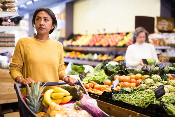 Image Pensive latina buying groceries at store, walking with shopping cart among shelves and choosing products