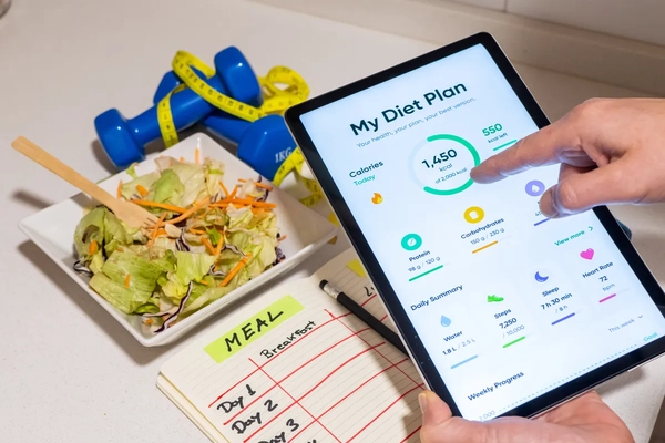 Hand interacting with tablet showing a personalized nutrition app calories, macronutrients and activity summary beside a fresh salad, dumbbells