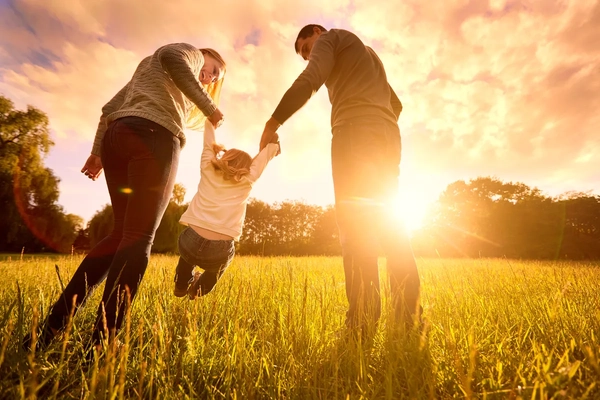 Image Parents hold toddler's arms, lifting them up over a field during sunset